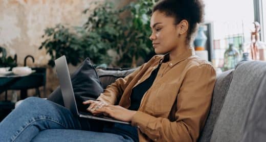 Une jeune femme est confortablement installée sur un canapé, travaillant sur un ordinateur portable, potentiellement en train de rédiger des mails de relance ou de postuler à des offres d'alternance.