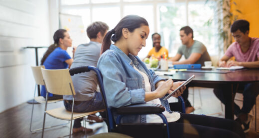 Jeune femme en fauteuil roulant (alternante RQTH) utilisant une tablette numérique lors d'une réunion professionnelle en entreprise. Illustration de l'inclusion et de la diversité au travail.